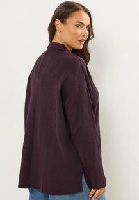 Overmaatse, dieppaarse cardigan van een textuurrijke, geribbelde stof. Bevat een kraag en zijsplitten, gecombineerd met lichtblauwe denim.