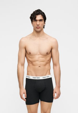 Jeune homme aux cheveux bouclés foncés portant un boxer noir avec une ceinture blanche marquée "Organic Basics", debout devant un fond gris.