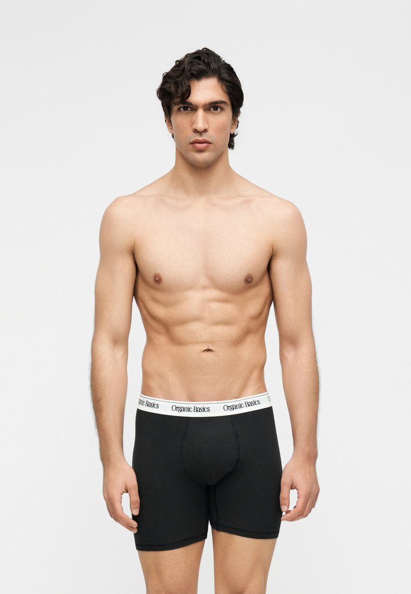 Νέος άνδρας με σκούρα, σγουρά μαλλιά, φοράει μαύρα μπόξερ με λευκή μέση που γράφει "Organic Basics" και στέκεται μπροστά σε γκρι φόντο.