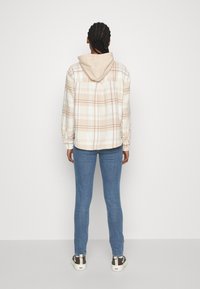 Camisa de franela a cuadros en beige y blanco con capucha, combinada con jeans azules. La camisa tiene puños abotonados y un corte relajado.