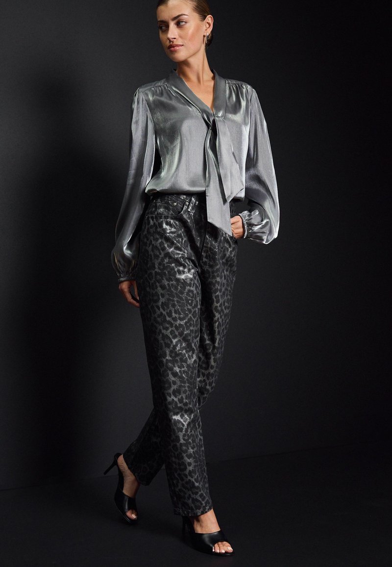 Bluză din satin argintie cu legătură frontală, pereche cu pantaloni din material leopard black și tocuri deschise negre, pe un fundal întunecat.