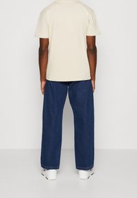 Osoba v béžové košili s krátkým rukávem, tmavě modrých volných džínech a bílých teniskách stojí zády k fotoaparátu na jednoduše pozadí.