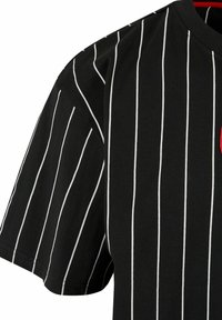 Cămașă neagră cu mânecă scurtă, evidențiată de dungi verticale albe, cu decolteu rotund și un detaliu subtil roșu, confecționată dintr-un material coton moale.