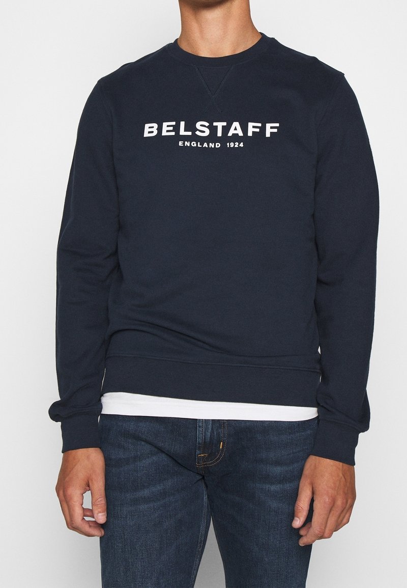 Homme portant un sweat-shirt bleu marine avec un texte blanc "BELSTAFF ENGLAND 1924", un jean bleu foncé et une chemise blanche qui dépasse de l’ourlet.