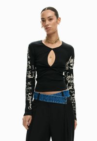 Zwart longsleeve top met sleuteloogdesign, met gestructureerde stof en witte handgeschreven patronen op de mouwen; gecombineerd met een blauwe denimriem.