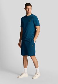 Vyriškas sportinis komplektas, apimantis žalią apvalią kaklo marškinėlį ir atitinkančius šortus. Abu turi tekstūruotą apdailą ir mažą geltoną logotipo detalę.