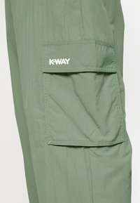 Zelená, lehká látka s vrásčitou texturou. Obsahuje klopovou kapsu a logo K-WAY v bílé barvě na přední kapse. Svislé švy jsou přítomny.