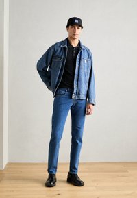 Jeansjakke med sidelommer, sort polo-shirt, blå jeans og sorte sko. Kasketten har et logo-patch; modellen står mod en neutral baggrund.