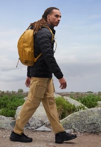 Black quilted jacket, beige cargo pants with multiple pockets, yellow backpack, and black sneakers. Outdoor setting with rocks and greenery.