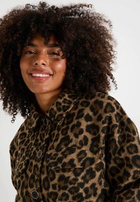 Mujer sonriente con cabello rizado, vistiendo una chaqueta abotonada de estampado de leopardo marrón y negro, contra un fondo liso.