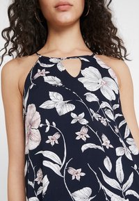 Marineblå blommig halternecktop med stora rosa och vita blommönster, nyckelhålsdetalj vid halslinningen och lätt tygstruktur.