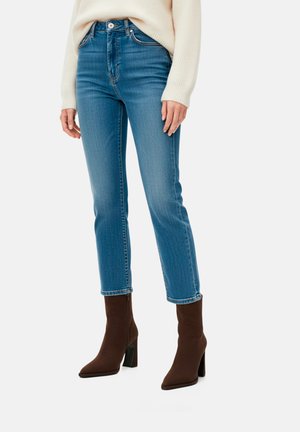 Donna con jeans blu corti, maglione crema e stivaletti marroni con tacco alto, in piedi su uno sfondo bianco.
