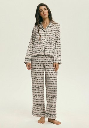 Kobieta stojąca boso, ubrana w beżowo-czarny, wzorzysty komplet piżamy z długim rękawem, z zapinanym na guziki topem i szerokimi spodniami.