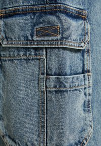 Denimtyg i ljusblått med kontrasterande orangea sömmar, en ficka med en rektangulär etikett och en texturerad yta.