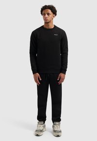 Zwarte sweatshirt met ronde hals en geborduurd logo, gecombineerd met zwarte joggerbroeken en grijze sneakers. Zachte textuur, casual ontwerp.