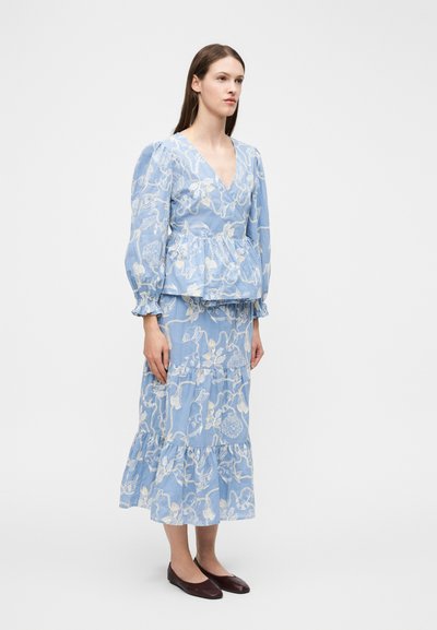 Kvinde stående iført en blå bluse med lange ærmer og et lagdelt skørt med hvide blomsterprints, kombineret med mørkebrune flade sko.