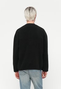 Svart fleece-tröja med en avslappnad passform, sjunkna axlar och en horisontell söm över ryggen. Kombinerad med ljusblå denimjeans.