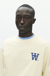 Jeune homme aux cheveux courts portant un sweat-shirt crème avec un logo "W" bleu et un sous-pull bleu clair sur un fond blanc uni.