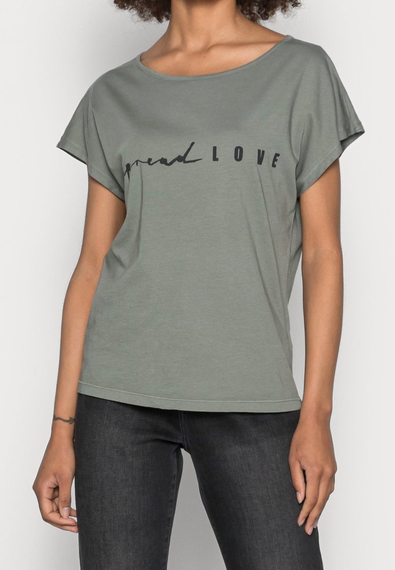 Femme portant un t-shirt vert à manches courtes avec le texte "spread LOVE" et un jean noir, debout avec les bras détendus le long du corps.