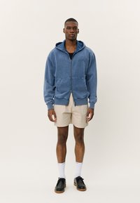 Blauwe rits hoodie van zacht materiaal, met een voorzak en een verstelbare capuchon, gecombineerd met beige shorts en zwarte schoenen.