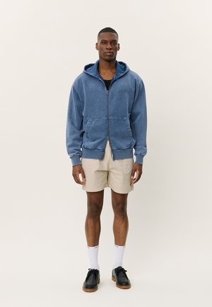 Veste zippée bleue en tissu doux, dotée d'une poche frontale et d'une capuche réglable, associée à un short beige et des chaussures noires.