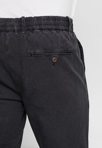 Zwarte stoffen broek met een elastische tailleband en een achterzak met een bronzen knop. De textuur is glad met een subtiel patroon.