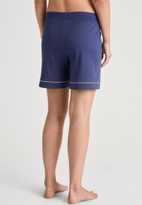 Marineblauwe katoenen shorts met een zachte textuur, elastische tailleband en witte biezen langs de zoom. Zicht van achteren, blote voeten zichtbaar.