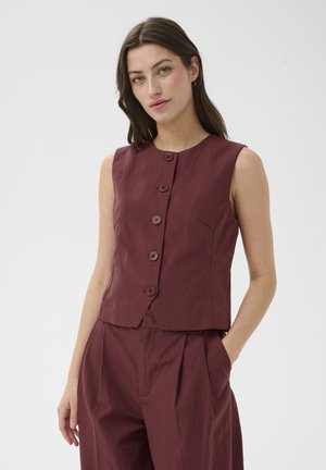 Femme aux longs cheveux foncés portant un gilet bordeaux sans manches boutonné et un pantalon assorti, debout avec une main dans la poche sur fond blanc.