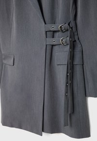 Blazer gris ajusté avec deux sangles horizontales et boucles en argent sur le devant, présentant des poches à rabat et un design structuré.