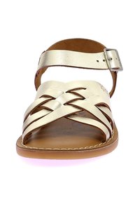 Sandalia de cuero dorado con correas entrelazadas, hebilla ajustable en el tobillo y plantilla de cuero marrón. Suela plana con detalles de costura sutil.
