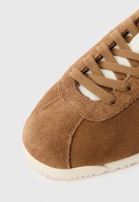 Brown suede sneaker featuring a rounded toe, cream laces, and a textured rubber sole with a wavy pattern.