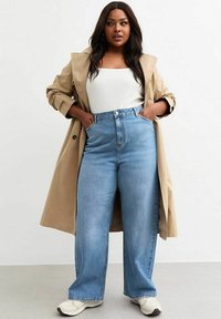 Trench coat beige sobre una camiseta de tirantes blanca con canalé, combinado con jeans de tiro alto y pierna ancha de color azul claro y zapatillas color crema.