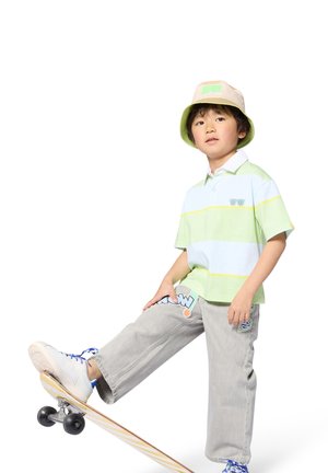 Lichtgroen-wit gestreept poloshirt, grijze broek met kleurrijke patches, witte sneakers en een beige bucket hat met groene accenten.