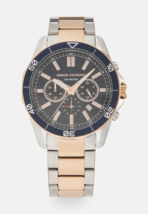 Zegarek chronograf z ciemną tarczą, srebrnym i różowym złotym stalowym bransoletą, sześciokątną lunetą oraz trzema podtarczycami. Wodoszczelny do 100 metrów.