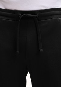 Black sweatpants with a visible drawstring waistband tied in a bow, worn with a white shirt tucked in.