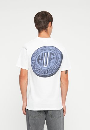 Hvid bomulds t-shirt med et stort, rundt marineblåt grafisk print med "HUF" tekst og detaljeret design på bagsiden. Grå jeans vist.