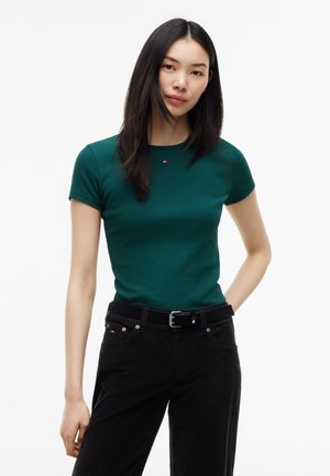 Frau mit glatten schwarzen Haaren, die ein türkisfarbenes kurzärmliges Shirt und schwarze Hose mit schwarzem Gürtel trägt, steht vor einem schlichten weißen Hintergrund.