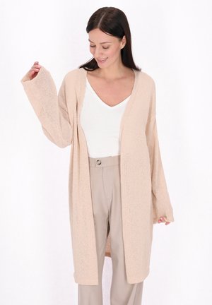 Beige gestrickte Cardigan mit lockerer Passform, offenem Frontbereich und langen Ärmeln, kombiniert mit einem weißen V-Ausschnitt-Shirt und beigen Hosen.