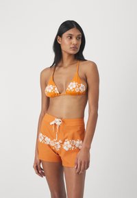 Haut de bikini orange avec un motif floral blanc et des bretelles réglables, associé à un short orange assorti présentant un design floral similaire et un cordon de serrage.