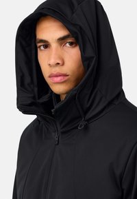 Melnā kapuci jaka, izgatavota no gludas, matētas auduma. Aprīkota ar rāvējslēdzēja aizdari un regulējamiem aukliņām uz kapuces. Nav redzamu rakstu vai akcentu.