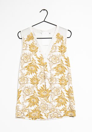 Ærmeløs top med V-udskæring, lavet af hvidt stof dekoreret med gule blomsterdesigns og grønne detaljer, hængende på en sort bøjle.