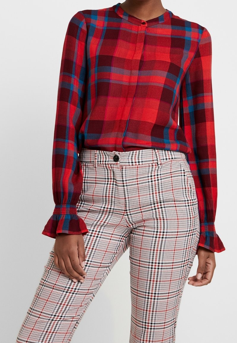 Blouse à manches longues rouge et bleue à carreaux avec un col rond. Associée à un pantalon à carreaux noir et blanc taille haute avec des détails de volants au niveau des poignets.