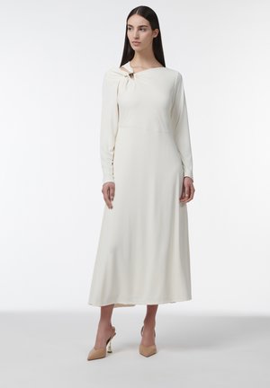 Vrouw met lang donker haar die een crèmekleurige midi-jurk met lange mouwen en een uitsnijding op de schouder draagt, gecombineerd met beige hoge hakken, staand tegen een witte achtergrond.