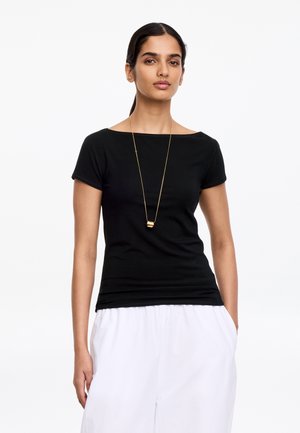 Kvinde iført en sort kortærmet top, langt gyldent halskæde og hvide bukser, stående med hænderne i lommerne på en hvid baggrund.