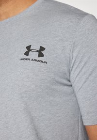 Acercamiento de una persona que lleva una camiseta de rayón de color gris claro de Under Armour, con el logotipo y el nombre de la marca en negro en el pecho.