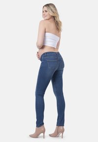 Jeans skinny de maternidade em denim azul escuro com uma textura suave. Apresenta uma cintura média e bolsos traseiros. Usados com um top branco sem alças.