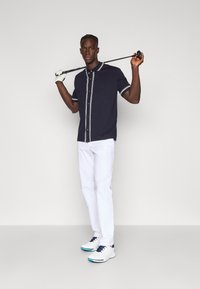 Camisa polo azul-marinho com botões brancos à frente e detalhes listrados nas mangas, combinada com calças brancas e sapatos de golfe brancos, segurando um taco de golfe.