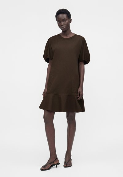 Γυναίκα που φοράει ένα σκούρο καφέ φόρεμα μέχρι το γόνατο με φουσκωτά μανίκια και φουσκωτή ζώνη, συνδυασμένο με μαύρα σανδάλια με χαμηλό τακούνι.