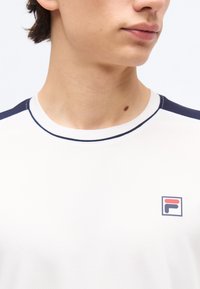 Νεαρός που φοράει λευκό κοντομάνικο μπλουζάκι με σκούρο μπλε διακοσμητική λεπτομέρεια και μικρό κόκκινο και σκούρο μπλε λογότυπο Fila στο στήθος.