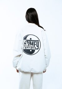 Biała, oversize'owa kurtka z czarnym graphic yin-yang i napisem "GRIMEY" umieszczonym na środku pleców, charakteryzująca się gładką, matową teksturą.